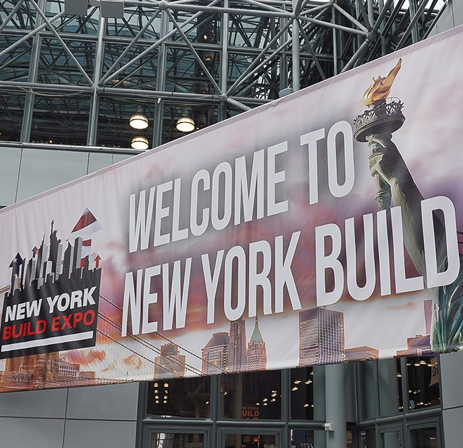 Banner reading 'Welcome to New York Build Expo' hanging inside a glass-roofed venue with skyline graphics in the lower portion and the Statue of Liberty torch image on the right side.