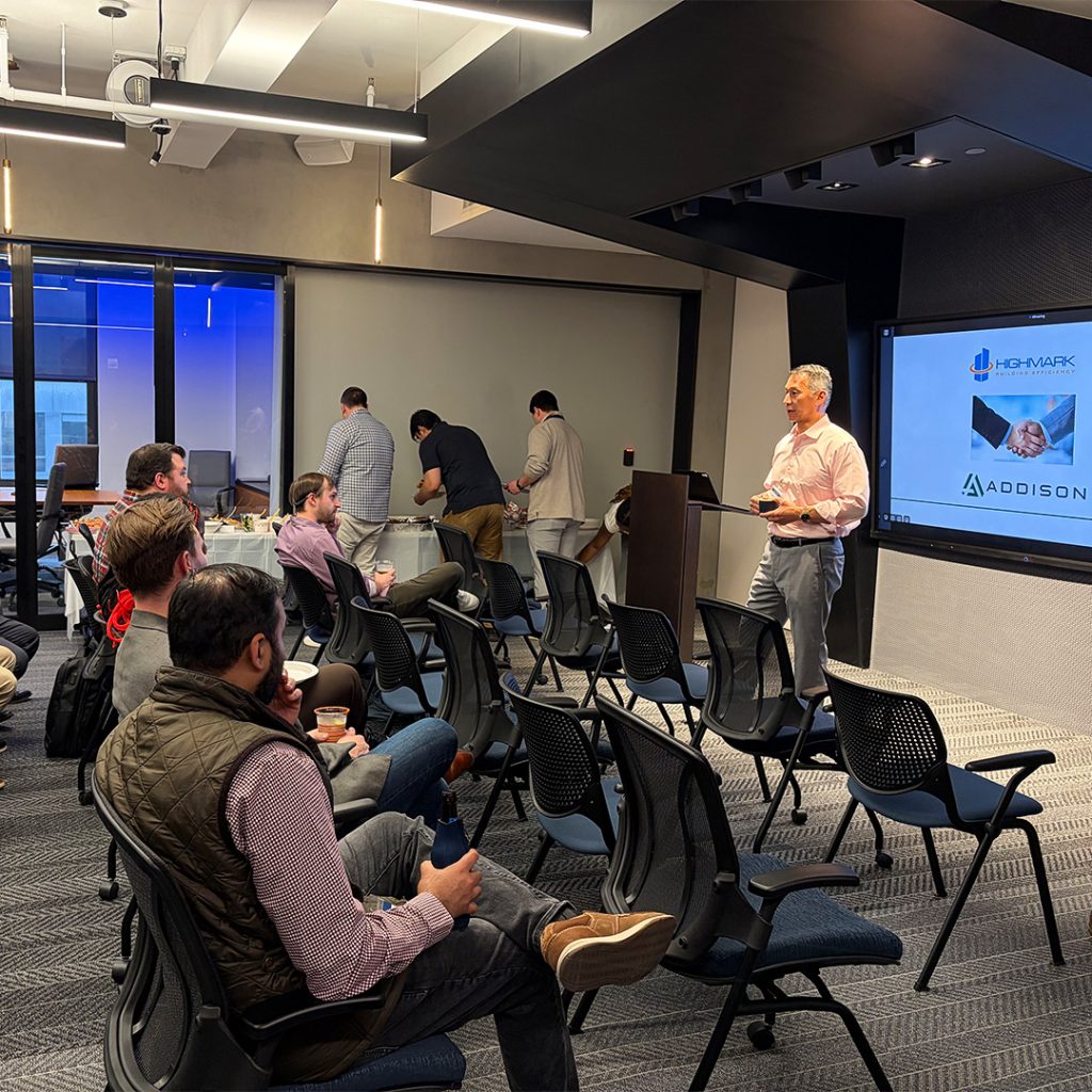 Man at a podium presenting to an audience in a modern conference room; large screen displays Highmark and Addison logos.