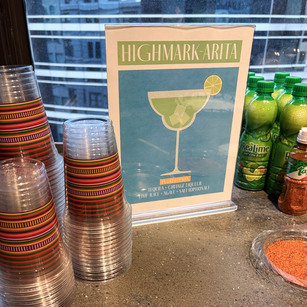 Poster reads 'HIGHMARK-ARITA' with a lime cocktail image; ingredients listed: tequila, orange liqueur, lime juice, agave, salt (optional). Nearby stacks of colorful plastic cups and lime juice bottles.